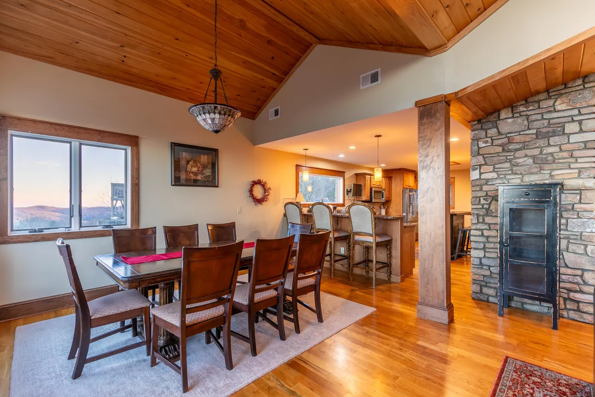 A cozy dining area with a wooden table, set beside a striking stone fireplace. Large windows reveal scenic outdoor views while the open kitchen area is visible in the background, emphasizing the home’s open-concept design.