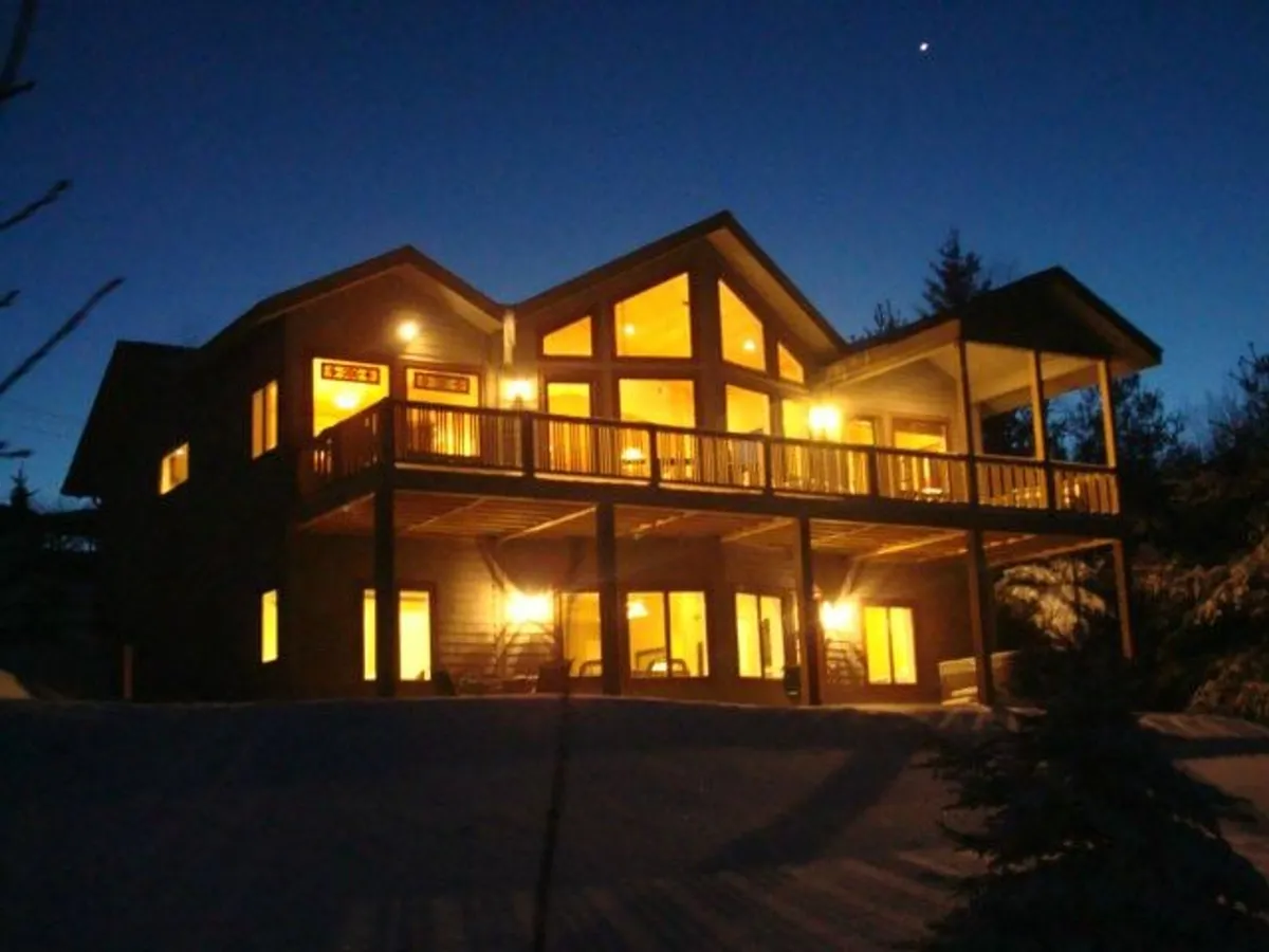 The exterior of a spacious mountain home with expansive windows and a welcoming balcony, softly illuminated by outdoor lighting. The scene exudes warmth, inviting guests to enjoy sunset views, gather under the stars, and relax in a cozy, elevated setting surrounded by the natural beauty of Blowing Rock.