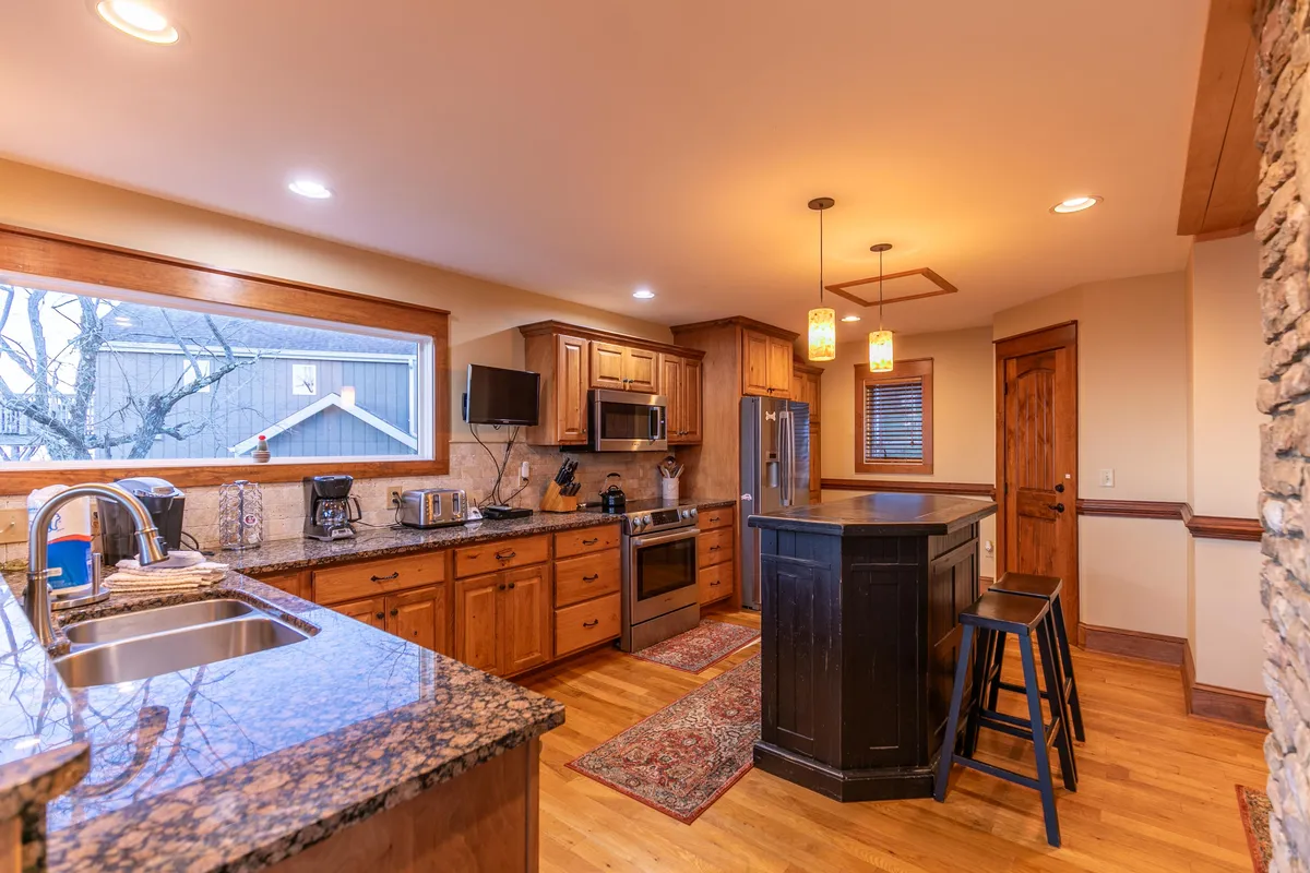 A bright, inviting kitchen with granite countertops, sturdy wooden cabinets, an island bar with stools for casual dining, and a large window that lets in natural light. The space is clearly designed for both meal preparation and socializing.