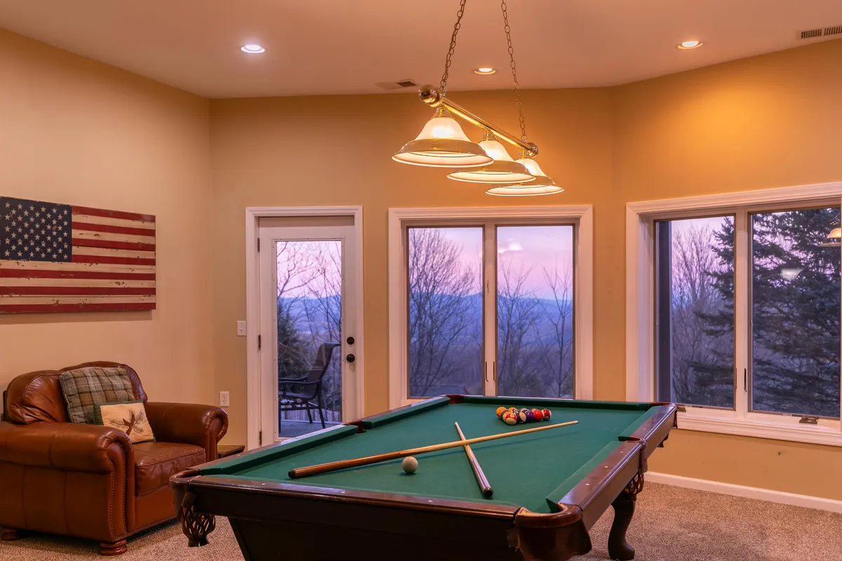 This interior image features a recreation room with a pool table, leather couch, and large windows framing the outdoors. The space is designed for group activities like game nights, friendly competition, and cozy conversations, all while connecting visually with the scenic surroundings.