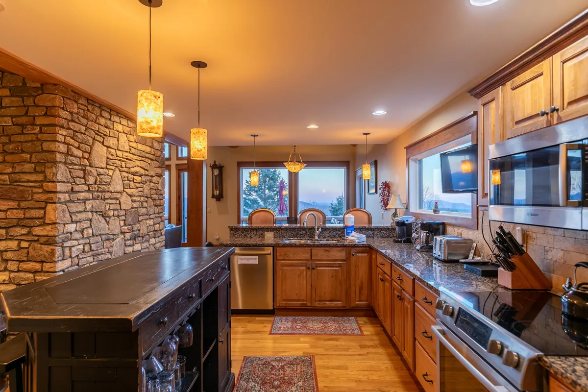 A warm, sophisticated gourmet kitchen with granite countertops, stone accents, pendant lighting, and large windows. The kitchen is both functional and stylish, ideal for chef-prepared breakfasts, wine tastings, or simply admiring the view while cooking or entertaining.