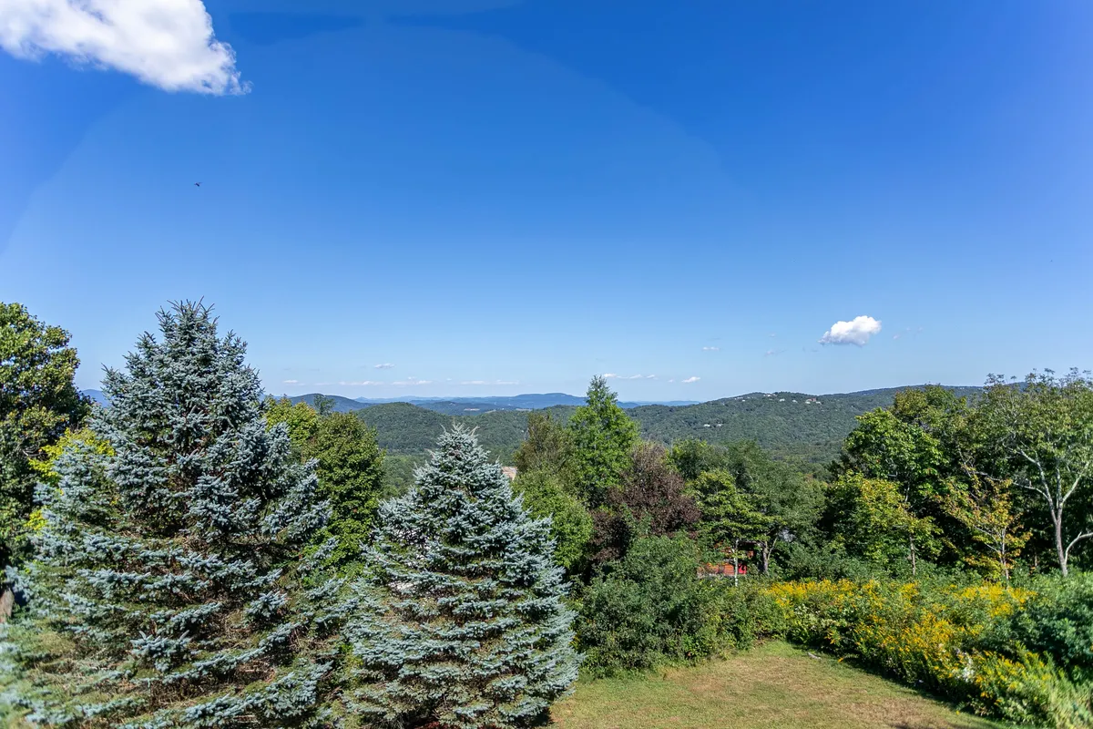 A classic mountain vista unfolds with lush greenery in the foreground and distant blue mountains under a wide, cloud-streaked sky. The scene is peaceful, hinting at miles of hiking trails and nature experiences just beyond the home.