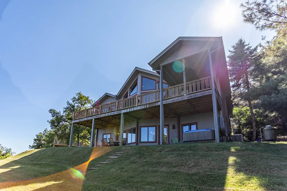 A wide view of a spacious, partially covered deck attached to a modern mountain home. The deck features plenty of open space for outdoor gatherings, surrounded by landscaped yard and tall trees. The architecture provides both exposed and sheltered areas, ideal for all-season enjoyment.