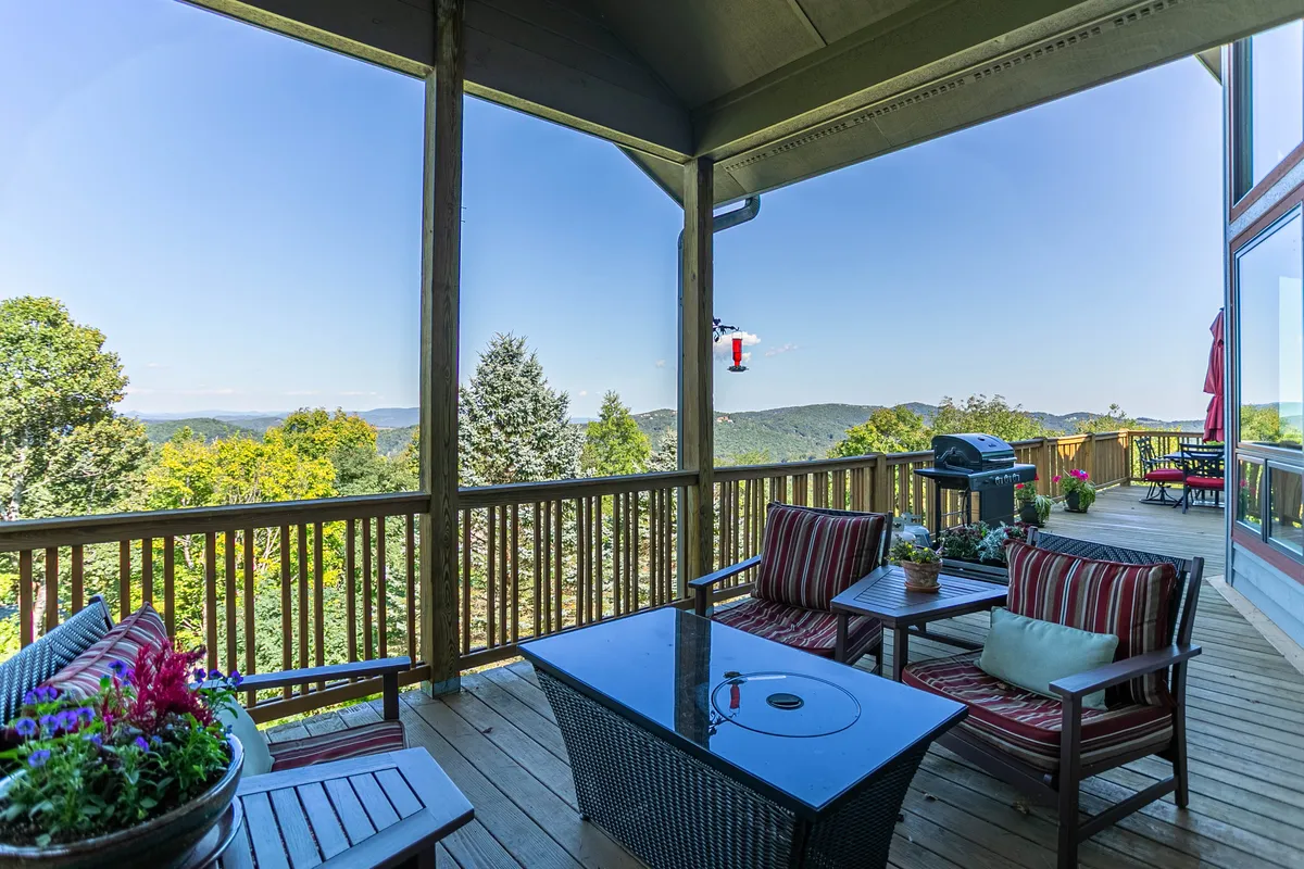 The image showcases a sweeping view of the Blue Ridge Mountains from a well-furnished outdoor seating area. The deck features comfortable chairs, a grill, and vibrant flower pots, all set against a backdrop of sunset hues and lush greenery.