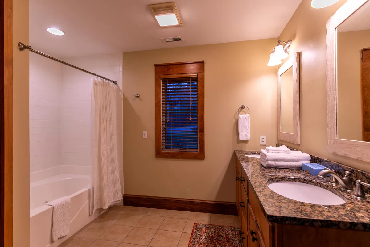 A tranquil, spa-like bathroom featuring a deep soaking bathtub, granite countertop, and a large mirror. Gentle natural light comes in through a window with blinds, creating a soothing space to relax and recharge.
